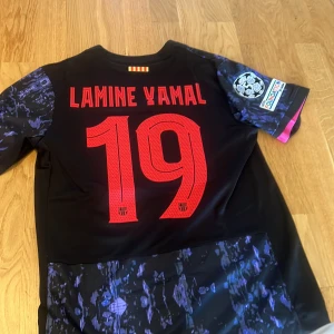 FC Barcelona Lamine Yamal Nike tröja - Säljer en FC Barcelona matchtröja med Lamine Yamal 19 på ryggen. Tröjan är svart med lila och rosa mönster, rosa detaljer och klubbmärke, samt Champions League och UEFA Foundation badges på ärmen. Tillverkad i lätt och ventilerande material. 
