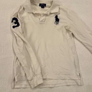 Vit långärmad pikétröja Ralph Lauren - Vit långärmad pikétröja från Ralph Lauren med klassisk krage, knäppning och stor marinblå polospelare broderad på bröstet. Snygg detalj med siffran 3 på ärmen. Tröjan är i bomull och har en relaxed fit som passar perfekt till jeans. Litet hål bak i nacken som dock inte syns under kragen  