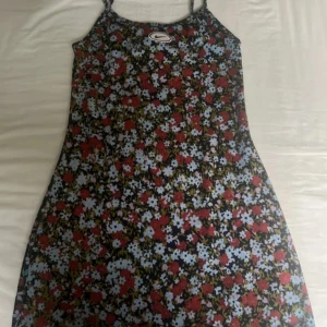 Blommig kortklänning från Nike XS - Supersöt kortklänning från Nike med färgglatt blommönster i rött, blått, lila och vitt på svart botten. Smala axelband och figurnära passform. Perfekt för dig som vill sticka ut med en sportig och lekfull vibe. Materialet är mjukt och stretchigt bomullstyg.