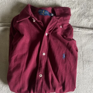 Vinröd skjorta från Ralph Lauren M - Vinröd skjorta från Ralph Lauren i bomullsmaterial, klassisk krage och knäppning framtill. Broderad blå logga på bröstet och vita knappar. Featherweight mesh-tyg som känns lätt och skönt. Perfekt för dig som gillar stilrena plagg med premiumkänsla.