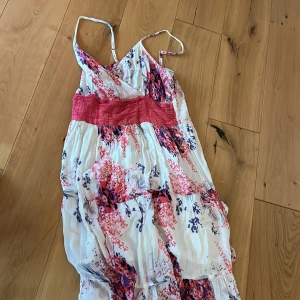 Blommig sommarklänning från Intuition - Söt långklänning från Intuition med blommigt mönster i rosa, rött och blått på vit botten. Klänningen har smala axelband, omlottliv och en bred rosa midjedetalj. Perfekt för varma dagar och har en luftig, flowy känsla.