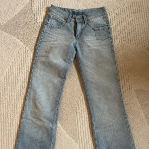 Ljusblå lågmidjade jeans med rhinestones - Snygga ljusblå jeans med raka ben och coola rhinestones på fickorna fram och bak. Jeansen har klassisk femficksdesign, bälteshällor och kontrastsömmar. Perfekta för dig som gillar en avslappnad men ändå trendig look.