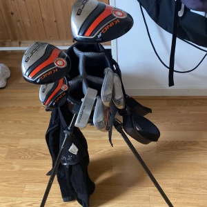 Cobra golfbag med klubbor - Säljer en sportig golfbag från Cobra i svart och grått med orange detaljer. Väskan har flera fack och praktiska bärremmar. Medföljer flera King JR golfklubbor, perfekt för dig som vill komma igång med golfen. Snygg och funktionell design för golfbanan.