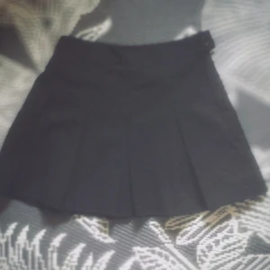 Svart plisserad kortkjol - Snygg svart kortkjol med plisserad design och knappdetalj i midjan. Kjolen har en klassisk och stilren look som är lätt att matcha med olika toppar och jackor. Perfekt för dig som gillar en tidlös vibe med lite edge.