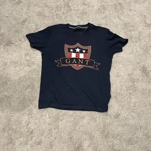 Mörkblå GANT t-shirt med tryck - Snygg mörkblå t-shirt från GANT med stort tryck framtill i form av ett sköldemblem med stjärnor och ränder i rött, vitt och guld. Klassisk rund halsringning och korta ärmar. Perfekt för en stilren och sportig look.