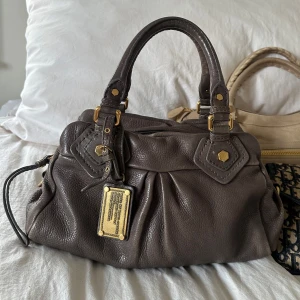 Mörkbeige grå Q Classic Baby Groove väska! - Köp helst via min vinted @juliavallberg 🤍Snygg beige/grå väska i fint skick från Marc by Marc Jacobs i äkta läder med gulddetaljer och dubbla handtag. Något slitet på lädret som syns på bilderna. Ger ett snyggt vintage intryck. Ren inuti och fint skick på detaljerna. Det är en otroligt välbevarad vintage modell! Perfekt för att ge en stilren touch till din outfit. Skriv privat för fler bilder. Mått 34x18x12 (undersida). Kan hämtas hemma hos mig i Nacka eller skickas men kan tyvärr inte mötas🤍
