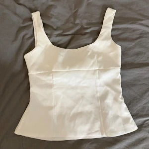 Vit linnetopp med rosett och cut out - Superfin vit linnetopp med breda axelband och en stor rosett baktill som ger en snygg cut out-detalj. Toppen har en lätt utsvängd passform och är tillverkad i ett slätt, mjukt material som känns lyxigt. Perfekt för att lyfta vilken outfit som helst!