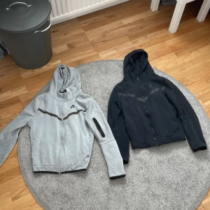 Nike zip hoodies med ficka på ärmen - Två snygga Nike hoodies med dragkedja och huva, en i ljusgrått och en i svart. Båda har en cool detalj med ficka på ärmen och en diskret rand över bröstet. Perfekta för chill dagar eller när du vill ha en sportig look. 1 för 400 men om du köper båda får du för 700 så du sparar 100kr