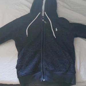 Svart hoodie från Polo Ralph Lauren - Svart hoodie från Polo Ralph Lauren med vit logga på bröstet. Tröjan har dragkedja, huva med vita snören och två fickor framtill. Perfekt för en chill och stilren look. Mjuk insida och klassisk passform.