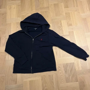 Marinblå Polo Ralph Lauren hoodie - Snygg marinblå hoodie från Polo Ralph Lauren med full zip och huva. Klassisk röd logga broderad på bröstet och två fickor framtill. Skulle passa dig runt 160-165 cm. Skriv om fler bilder eller funderingar.