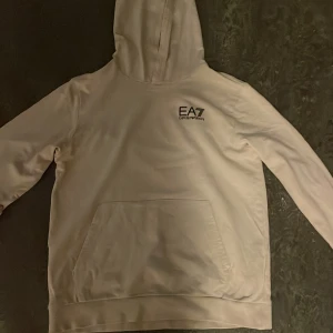 Bleach hoodie från Emporio Armani EA7 - Snygg vit hoodie från Emporio Armani EA7 med svart logga på bröstet. Klassisk känguruficka framtill och huva. Perfekt för dig som gillar stilrena och exklusiva plagg. Tillverkad i mjukt material som känns skönt mot huden.