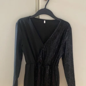 Svart glittrig omlottklänning S - Svart kortklänning med långa ärmar och omlott-skärning. Klänningen har glittriga paljetter framtill och en markerad midja. Perfekt för dig som vill sticka ut med en elegant och trendig look.