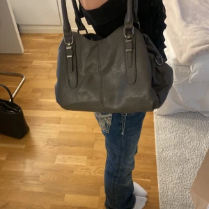 Mörkgrå axelväska i skinn - En mörkgrå praktisk väska att ha hängandes på axeln eller på armen. Några fickor och alla går att stänga till med antingen dragkedja eller en knapp. Ett band följer även med (bandet är i bra skick och samma färg som väskan. Har tidigare använts som en skol handväska och fungerar fortfarande toppen. Köpt på en marknad i Spanien.