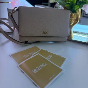 Beige axelväska från Michael Kors - Snygg beige axelväska från Michael Kors i äkta skinn med guldfärgade detaljer och MK-logga framtill. Väskan har justerbar axelrem, flera kortfack och dragkedjefack inuti. Perfekt storlek för det viktigaste och stilren design som lyfter varje outfit.