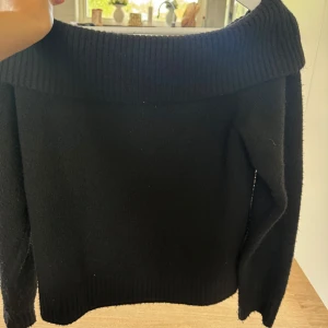 Svart offshoulder stickad tröja Neo Noir - Svart stickad tröja från Neo Noir med bred ribbad offshoulder-krage och långa ärmar. Tröjan har en klassisk och stilren look, perfekt för lager-på-lager och passar till många olika outfits.