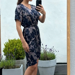 Mörkblå och beige mönstrad klänning - Snygg kortklänning med mörkblått och beige abstrakt blommönster. Klänningen har v-ringning, korta ärmar och knyt i midjan för en avslappnad passform. Tillverkad i ett lätt och mjukt material som känns skönt mot huden.