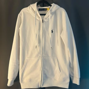 Vit hoodie från Polo Ralph Lauren XL - Vit hoodie från Polo Ralph Lauren i storlek XL. Klassisk design med dragkedja, huva med snörning och två fickor framtill. Broderad marinblå logga på bröstet. Mjuk bomullskänsla och ribbade muddar för extra komfort.