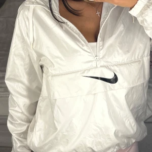 Vit Nike vindjacka - Snygg vit vindjacka från Nike med stor svart logga på ryggen och mindre logga framtill. Jackan har huva, halv dragkedja och stor ficka med dragkedja på magen. Superfint skick!