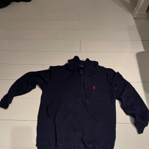 Mörkblå hoodie från Polo Ralph Lauren - Snygg mörkblå hoodie från Polo Ralph Lauren med dragkedja och huva. Klassisk röd logga broderad på bröstet, två fickor framtill och justerbara snören i huvan. Perfekt för en chill och stilren look.