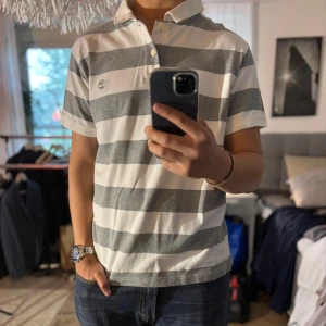 Randig pikétröja från Timberland - Snygg pikétröja från Timberland i storlek L med breda grå och vita ränder. Klassisk krage, två knappar och korta ärmar. Tröjan är i bomull och har Timberland-loggan broderad på bröstet. Perfekt för en chill och stilren look.