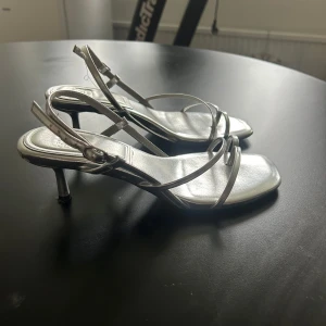 Silvriga klackskor - Silvriga klackar från ASOS med smala remmar och låg klack. Justerbart spänne vid ankeln. Säljer då dom inte kommer till användning, använt en gång endast.