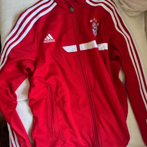 Röd Adidas Bayern München jacka - Säljer en röd Adidas Bayern München vindjacka med vita detaljer och klassiska tre ränder längs ärmarna. Jackan har dragkedja, hög krage och klubbmärke på bröstet. Perfekt för dig som vill ha sportig stil och visa vilket lag du hejar på!