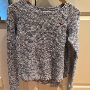 Blåvit stickad tröja från Hollister - Säljer en blåvit stickad tröja från Hollister med lång ärm och rund hals. Tröjan har ett melerat mönster och Hollisters klassiska logga broderad på bröstet. Perfekt för lager-på-lager och chill dagar.