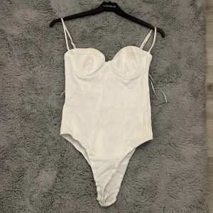 Vit baddräkt med bygel och smala band - Supersnygg vit body med bygelkupor och smala axelband. Stilren och enkel design som ger en clean look. Materialet har en lätt glans och känns mjukt mot huden. Perfekt för dig som gillar minimalistisk stil.