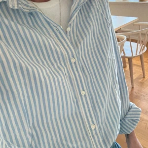 Blåvit randig skjorta  - Snygg skjorta i blåvitt randigt från H&M Klassisk krage, långa ärmar och knäppning framtill. Skjortan är i mjuk bomull och har en avslappnad passform, perfekt att bära öppen över ett linne eller knäppt till jeans.