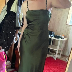 Mörkgrön långklänning i satin - Snygg mörkgrön långklänning i satin med smala axelband dekorerade med stenar. Klänningen har rak halsringning och en figurnära passform som ger en elegant siluett. Perfekt för dig som vill sticka ut med en stilren look.