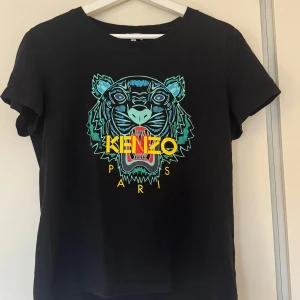 Svart Kenzo t-shirt Dam - Svart äkta t-shirt från Kenzo med ikoniskt tigertryck i blått, grönt och rött framtill. Stora gula Kenzo Paris-bokstäver över motivet. Nyskick. 