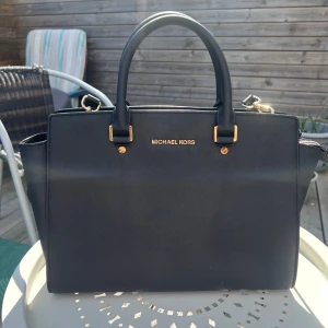 Svart handväska från Michael Kors - Snygg svart handväska från Michael Kors, modell Selma, med guldiga detaljer och logga framtill. Väskan har både handtag och justerbar axelrem, samt flera fack inuti och dragkedja med märkets logga. Stilren och rymlig modell i skinn som passar till allt. Mått: Längd i botten ca 32,5 cm. Längd bredaste parti upptill ca 40 cm. Höjd ca 25 cm.