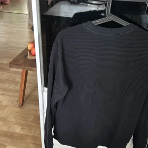 Svart slim fit tröja från Hugo Boss - Svart långärmad tröja från Hugo Boss med diskret broderad logga framtill. Slim fit-modell med ribbad krage och axelparti i lätt ribbat tyg. Perfekt för dig som gillar stilrena och klassiska plagg med en exklusiv touch. Storlek L barn men passar storlek S