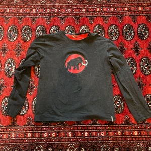 Svart långärmad tröja från Mammut - Svart långärmad tröja från Mammut med rött mammut-tryck på bröstet och röd logga längs ena ärmen. Tröjan har rund hals och röda detaljer på insidan av kragen. Perfekt för dig som gillar outdoor och streetwear vibes.