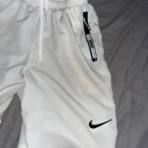 Vita Nike shorts med dragkedja - Snygga vita shorts från Nike med svart logga på benet och dragkedjeförsedda fickor. De har elastisk midja med snörning för skön passform och är perfekta för sport eller chill. Materialet känns lätt och andas bra.    Helt nya kar nog kvitto kvar