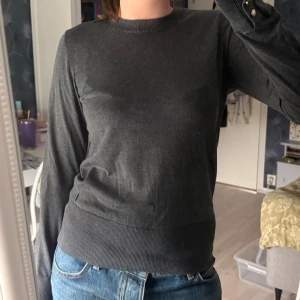 Mörkgrå stickad tröja Holly & Whyte - Mörkgrå stickad tröja från Holly & Whyte med rund hals och långa ärmar. Guldbricka har lossnat lite