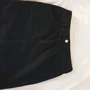 Svart jeanskjol från &Denim, strl 38 - Svart kort jeanskjol från &Denim i storlek 38. Klassisk femficksmodell med rå kant nertill och bälteshällor. Kjolen har knapp och dragkedja framtill samt två bakfickor. Perfekt till sneakers eller boots.