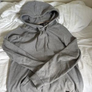 Grå hoodie från BOSS Homewear - Snygg grå hoodie från BOSS Homewear med klassisk huva och snörning. Tröjan har långa ärmar och en diskret metallogo vid nederkanten. Tillverkad i mjuk bomullsmix som är perfekt för chill dagar eller när du vill ha en clean look.