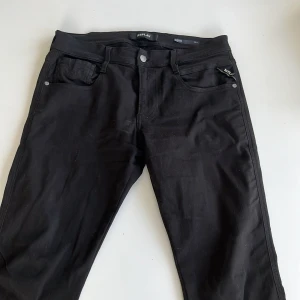 Svarta Replay Anbass jeans - Svarta jeans från Replay, modell Anbass med slim fit. Klassisk femficksdesign, svarta sömmar och Replay Hyperflex-märke bak. Snygg, stilren look med stretchigt material som sitter tajt men bekvämt. Perfekta för dig som gillar en modern och clean stil.
