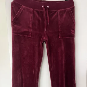 Vinröda velourbyxor Juicy Couture M - Vinröda mjukisbyxor från Juicy Couture. Storlek M. Använda men fortfarande fint skick! Metall ”knapparna” på snöret har tyvärr trillat av. Men annars inget fel på dem. Fickor med knappar baktill. Kan skicka fler bilder om så behövs. Står ej för frakten