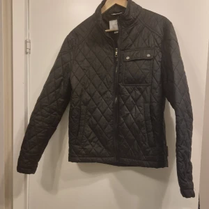 Svart quiltad jacka från Zara Man - Svart quiltad jacka från Zara Man i storlek M. Jackan har ett snyggt rutmönster, hög krage, dragkedja framtill och en bröstficka med knapp. Perfekt för dig som gillar stilren och modern look.