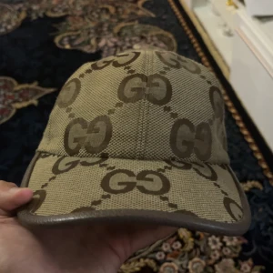 Beige keps från Gucci med GG-mönster - Snygg beige keps från Gucci med klassiskt GG-mönster i brunt över hela kepsen. Kepsen har böjd skärm och bruna kanter, tillverkad i ett slitstarkt tygmaterial. Perfekt för dig som vill sticka ut med en lyxig accessoar.