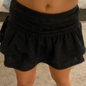 Svart volangkjol med shorts - Supersöt svart kortkjol med volanger och inbyggda shorts under. Kjolen har en bred, rynkad midja och dubbla lager för extra volym. Väldigt elastiskt. Tror den skulle passa M också. Använd fåtal gånger. Säljer en likadan i vit🥰