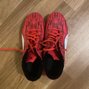 Röda Puma fotbollsskor strl 40 - Säljer ett par röda Puma fotbollsskor med svart och vitt mönster. Skorna har snörning, svart innersula och vit Puma-logga på sidan. Snyggt randigt mönster i svart på ovandelen och platt sula som passar för inomhusspel.