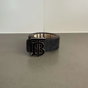 Svart Burberry bälte med monogramspänne - Snyggt svart bälte från Burberry med klassiskt rutmönster och stort metallspänne med Burberrys ikoniska TB-monogram. Bältet är gjort i skinn och har en exklusiv känsla med präglade detaljer längs remmen. Perfekt för att lyfta vilken outfit som helst.