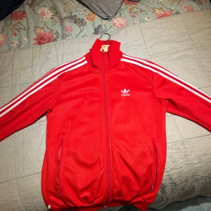 Röd Adidas track jacket med vita ränder - Röd Adidas track jacket med klassiska vita ränder längs ärmarna och vit logga på bröstet. Jackan har dragkedja framtill, hög krage och två fickor med dragkedja. Perfekt för en sportig och retro vibe.
