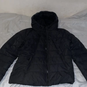 Svart pufferjacka från FB Sister XL - Svart pufferjacka från FB Sister i storlek XL. Jackan har huva med snörning, dragkedja framtill och quiltad design. Perfekt för kalla dagar och enkel att matcha med allt. Snygg och stilren look som funkar till alla outfits.