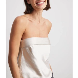  tubtopp i satin - Snygg axelbandslös tubtopp i champagnefärgad satin med bred kant upptill och elastisk baksida för perfekt passform. Toppen har en stilren och minimalistisk look som känns både lyxig och trendig. Perfekt att styla med jeans eller kjol för en cool vibe. Aldrig använd!