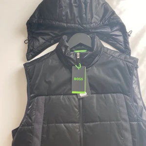 Svart dunväst från BOSS med huva. Stolek M  - Snygg svart dunväst från BOSS med avtagbar huva och dragkedja framtill. Västen har quiltad design, två dragkedjefickor och diskret BOSS-logga på ryggen. Perfekt för lager-på-lager och streetstyle. Materialet är glansigt och känns riktigt modernt.