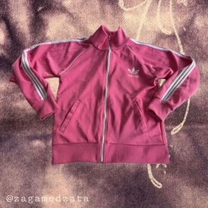 Rosa Adidas track jacket med stripes - Säljer en ikonisk rosa Adidas track jacket med vita stripes längs ärmarna och stor logga på ryggen. Jackan har dragkedja framtill, höga muddar och två fickor. Perfekt för dig som vill sticka ut med sportig retrostil.